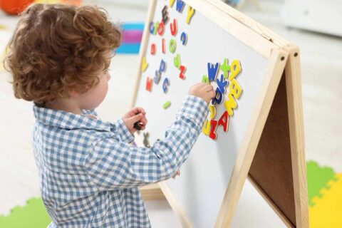 A young child places colorful magnetic letters on a wooden easel.