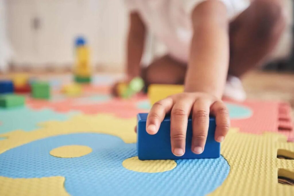 A child playing on a colorful foam mat holds a blue block.