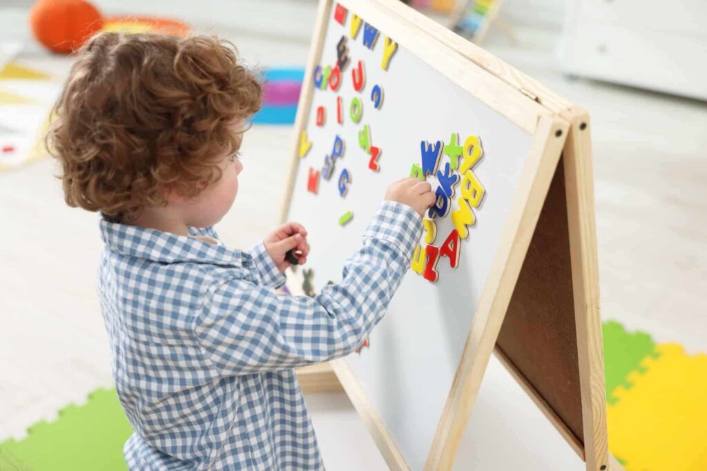 A young child places colorful magnetic letters on a wooden easel.