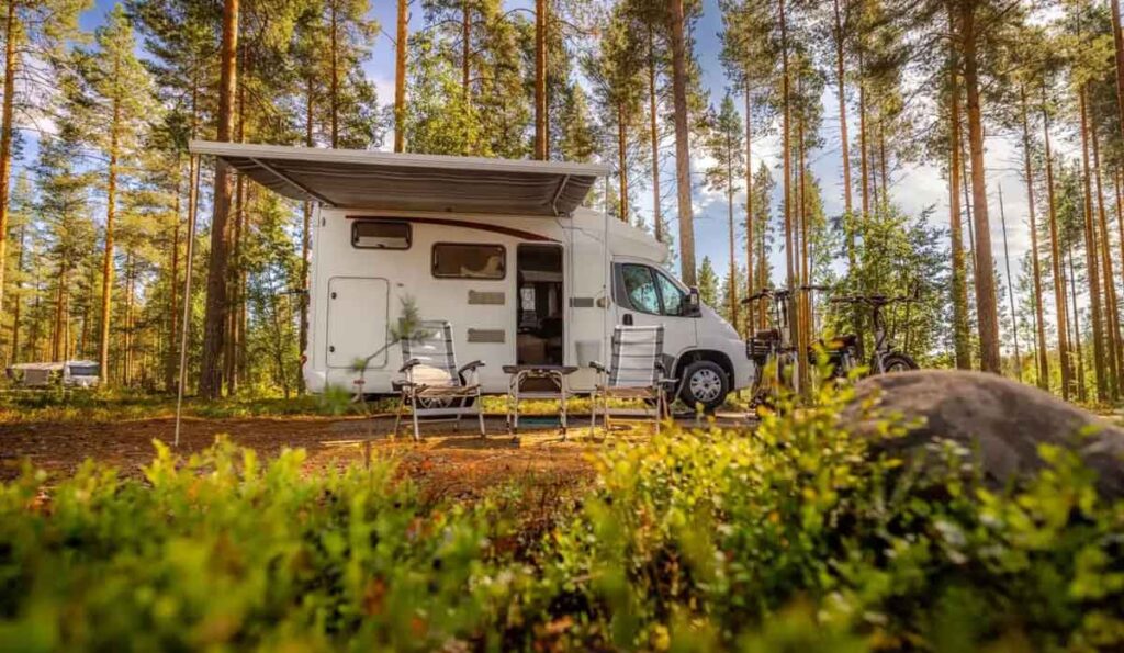Camper with an extended awning and chairs set up in a sunny forest. 