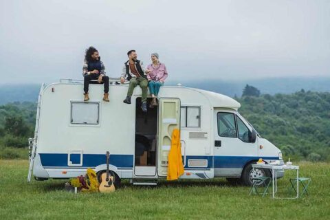 Three people sit on a camper roof in a lush field, smiling and chatting.