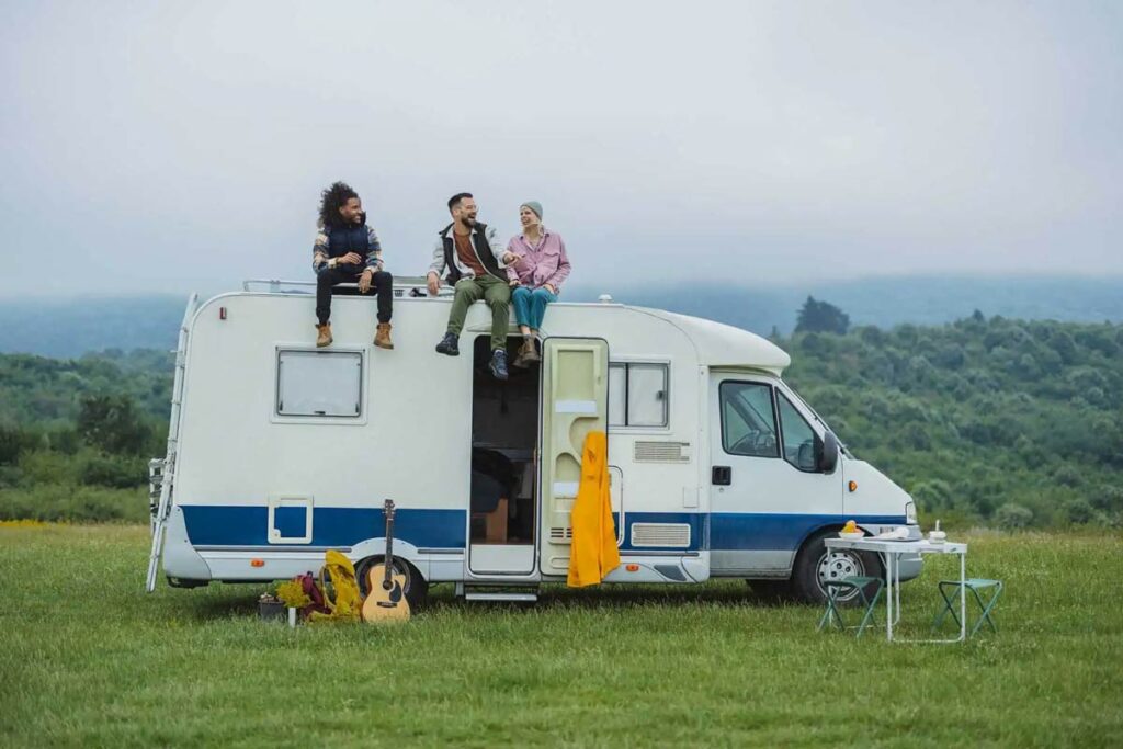 Three people sit on a camper roof in a lush field, smiling and chatting.