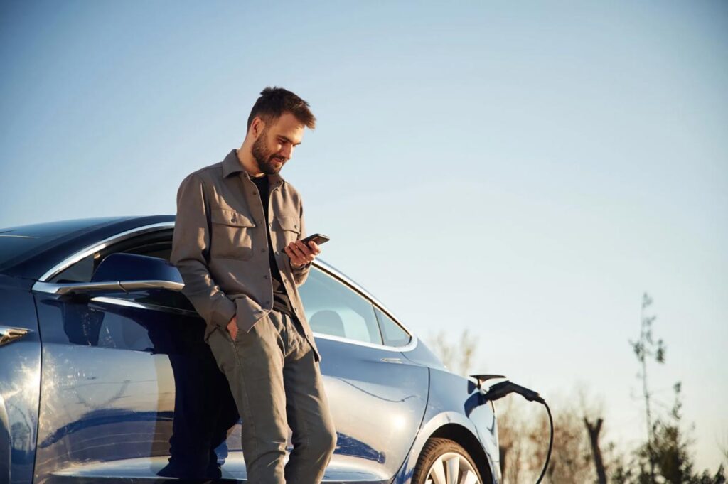 A man in casual attire leans on a charging blue electric car