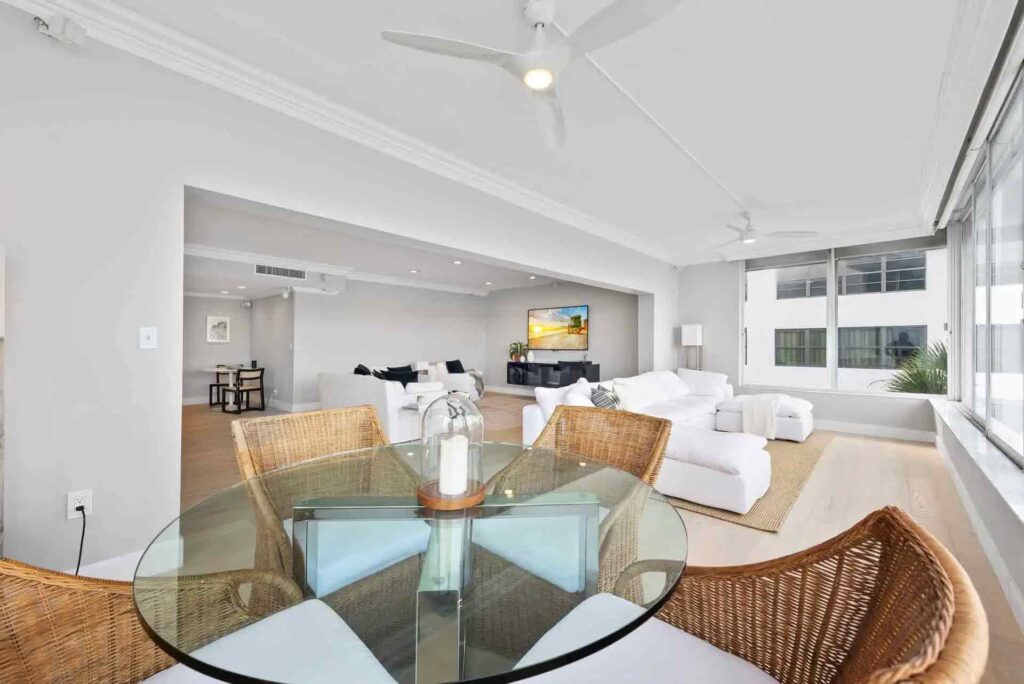 A modern living and dining area with white sofas, a glass-top table with wicker chairs, and large windows letting in natural light.