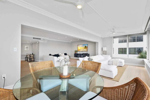A modern living and dining area with white sofas, a glass-top table with wicker chairs, and large windows letting in natural light.