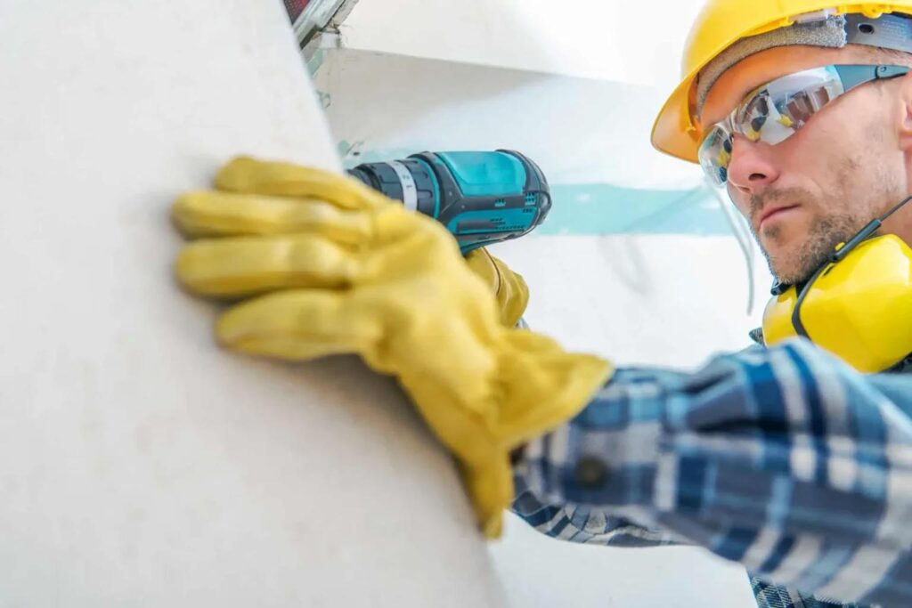 A focused construction worker in a yellow hard hat and gloves uses a power drill on a wall. 