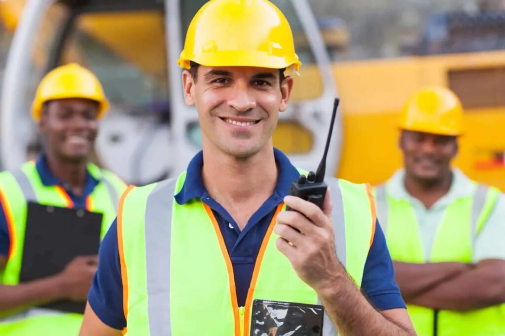 Smiling construction worker in a yellow hard hat and reflective vest holds a walkie-talkie. 
