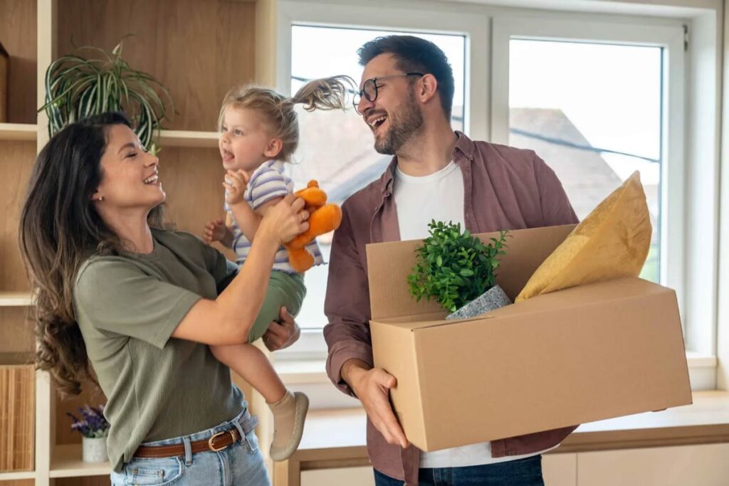 A joyful family scene with a mother holding a smiling toddler and a father carrying a cardboard box.