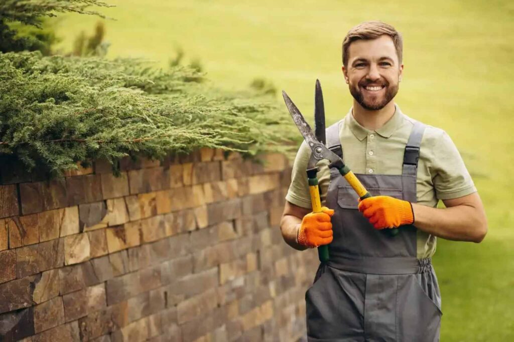 A smiling gardener stands outdoors holding pruning shears