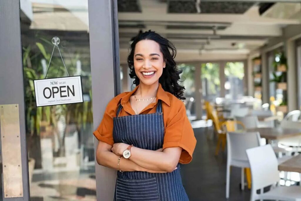 Smiling woman in an apron stands proudly at a café entrance with an Open sign. 